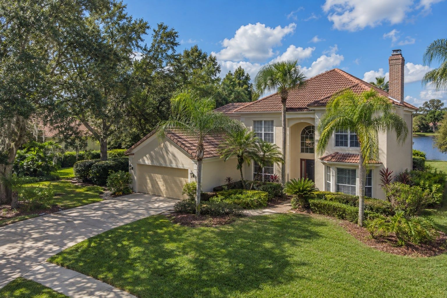 A two-storey stucco house with a tile roof and a brick chimney, seen from a low aerial angle among palms and live oaks under a blue sky